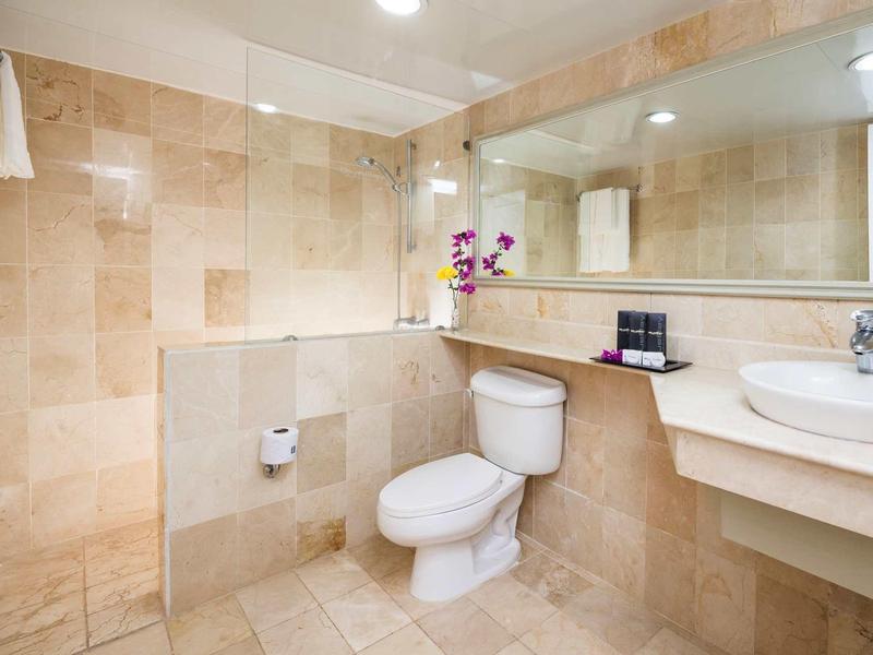 Helles Badezimmer mit beiger Fliesenwand, Toilette, Waschbecken, großem Spiegel und Dusche ohne Tür.