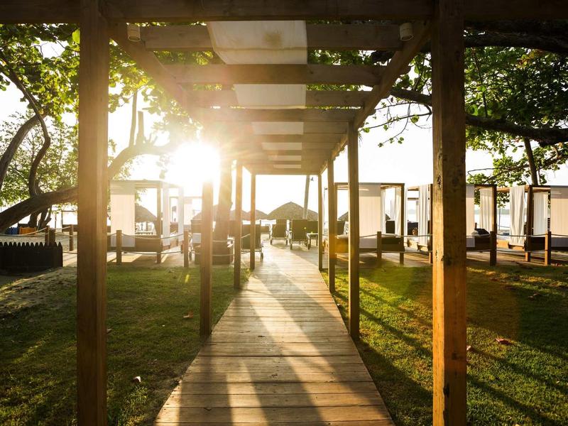 Sonnenlicht scheint durch Holzbalken über einem Weg auf eine grüne Wiese mit Liegestühlen.