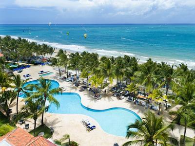 Strandresort mit geschwungenem Pool, Palmen, Liegestühlen und blauem Meer im Hintergrund.