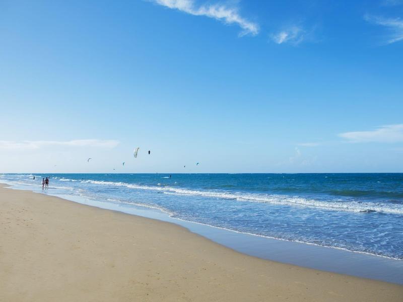 Weitläufiger Sandstrand mit sanften Wellen, blauem Himmel und vereinzelten Wolken am Horizont.