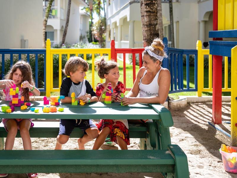 Vier Kinder sitzen an einem grünen Picknicktisch im bunten Außenspielbereich und spielen mit Bauklötzen.