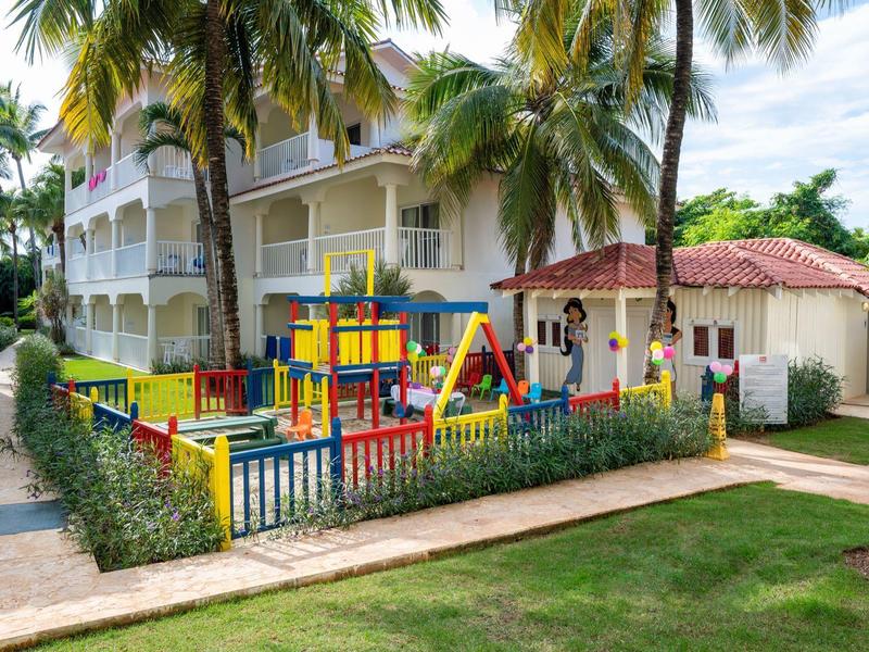 Bunter Kinderspielplatz mit Schaukeln und Rutsche, umgeben von Palmen vor einem Wohnhaus.