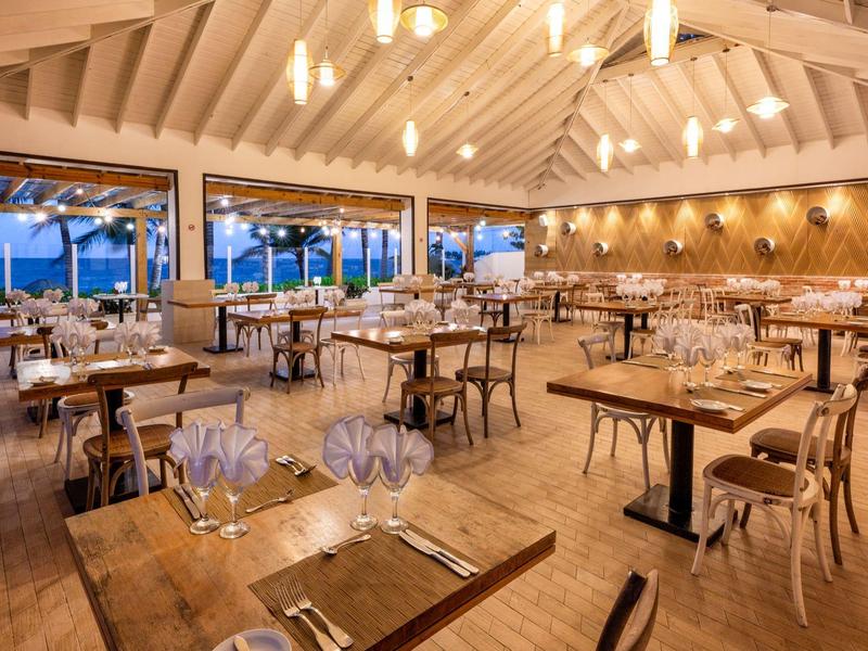 Helles Restaurant mit Holztischen, Stühlen, modernen Lampen und weitem Meerblick durch große Fenster.