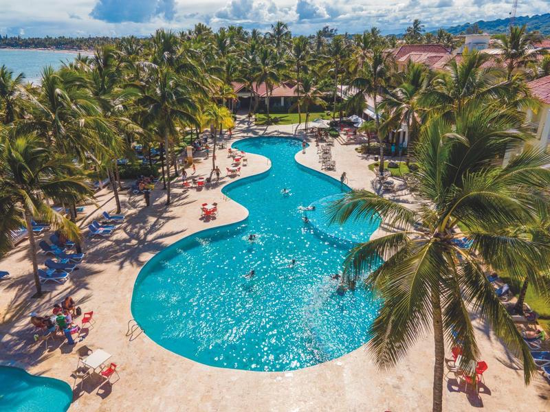 Großer, geschwungener Pool umgeben von Palmen und Liegestühlen in tropischer Resort-Umgebung.