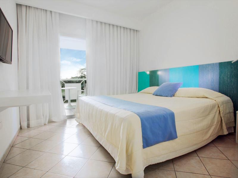 Modernes helles Schlafzimmer mit großem Bett, blau-grünem Kopfteil und Balkon mit Meerblick.