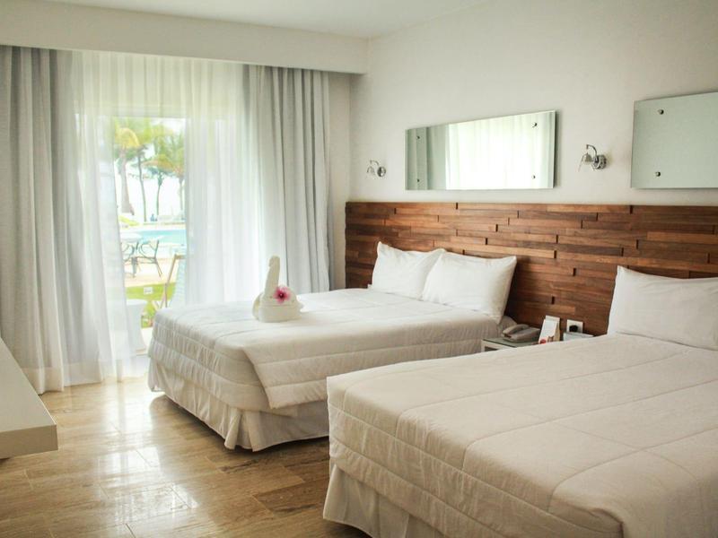 Helles Hotelzimmer mit zwei weißen Betten, Holzwand, Spiegeln und Blick auf Garten durch Vorhänge.