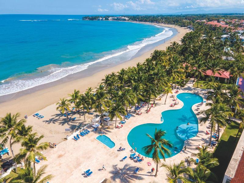 Langer Sandstrand, türkisblaues Meer, Palmen und großes, geschwungenes Schwimmbecken mit Liegestühlen.