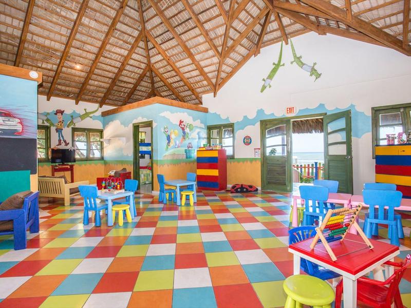 Bunter Kindergartenraum mit kleinem Mobiliar, blau-roten Tischen, gelben und blauen Stühlen, holzfarbenem Dach.