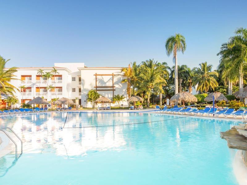 Großer Außenpool mit klar blauem Wasser, umgeben von Palmen und weißem Hotelgebäude im Sonnenschein.