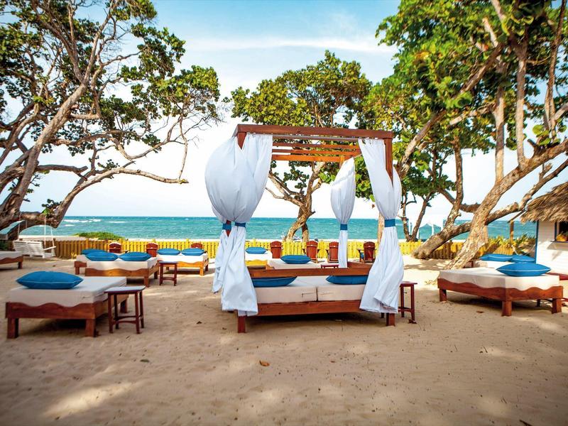 Strand mit Sand, Holzbetten, weißen Vorhängen und Bäumen mit Blick auf das blaue Meer.