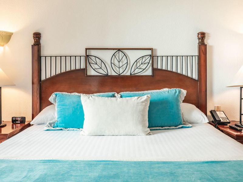 Modernes Doppelbett mit braunem Kopfteil, weißen und türkisfarbenen Kissen, Holztischen und Lampen.