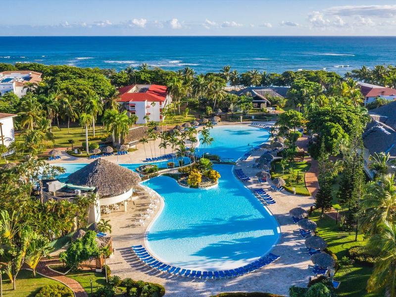 Großer blauer Pool mit Palmen und Liegestühlen, dahinter grüner Urwald und blauer Ozean unter blauem Himmel.