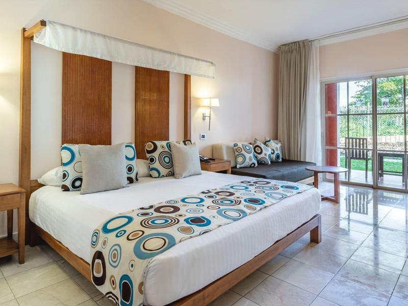 Helles Hotelzimmer mit Doppelbett, Holzakzenten, bunten Kissen und Balkon mit Gartenausblick.