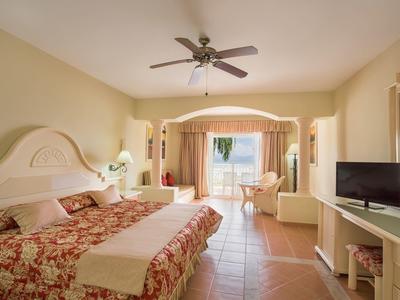 Hell eingerichtetes Hotelzimmer mit großem Bett, Ventilator, Fernseher und Balkon mit Blick nach draußen.