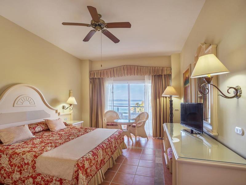 Gemütliches Schlafzimmer mit Doppelbett, Ventilator, Fernseher und Balkon mit Sitzgruppe.