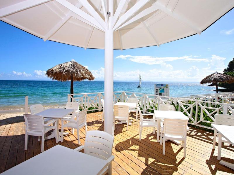 Sonnige Holzterrasse mit weißen Stühlen und Tischen, Meerblick und Sonnenschirmen am Strand.