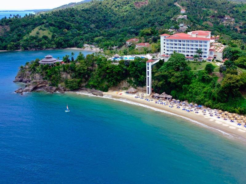 Bucht mit klarem blauem Wasser, Sandstrand, Hotel und grün bewaldetem Hügel im Hintergrund.