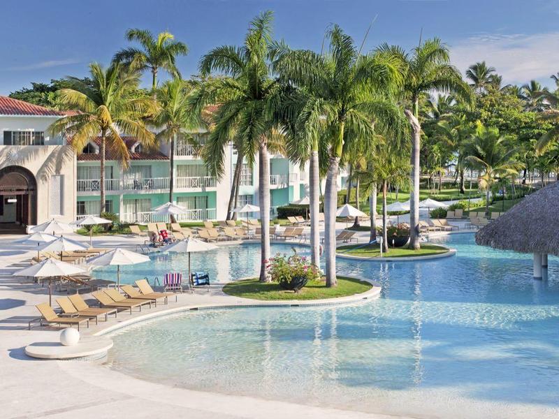 Großer Hotelpool mit Palmen, blauen Liegestühlen und hellem Gebäude im Hintergrund.