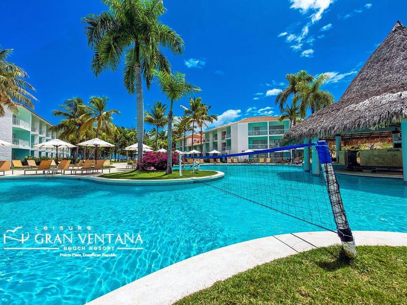 Großer Pool mit Volleyballnetz, umgeben von Palmen und mehrstöckigen Hotelgebäuden.