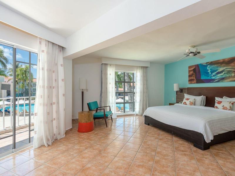 Helles Schlafzimmer mit großem Bett, türkisfarbener Wand, Fliesenboden und Blick auf den Pool.