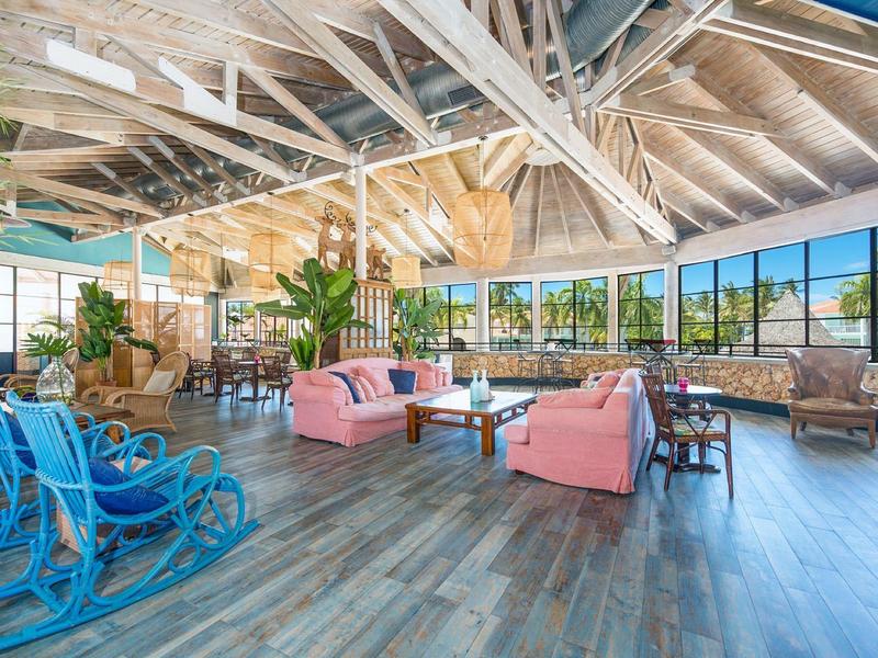 Großer heller Raum mit Holzbalkendecke, blauen Schaukelstühlen, rosa Sofas und vielen Pflanzen.