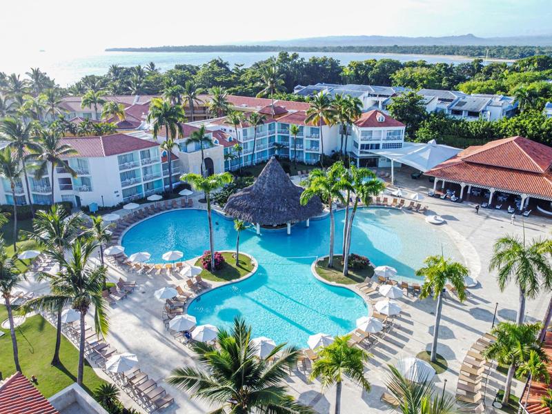 Große Hotelanlage mit großem Pool, Palmen, Sonnenschirmen und naher Meeresküste im Hintergrund.