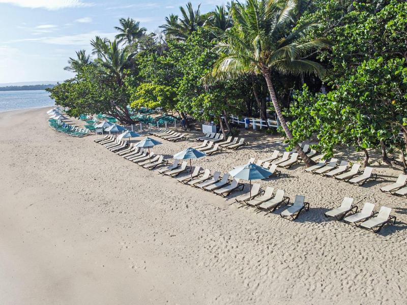 Palmen umgeben einen Sandstrand mit vielen weißen Liegestühlen in Reihen am Meer.