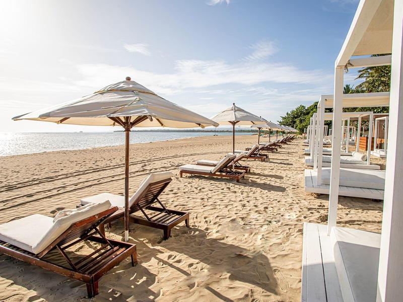 Strand mit Sand, mehreren Holzliegen und weißen Sonnenschirmen bei klarem Himmel.