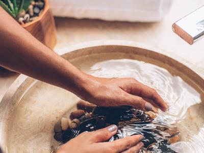 Hände tauchen in einen runden Steinwaschbecken mit Wasser und Steinen, Spa-Handtuch im Hintergrund