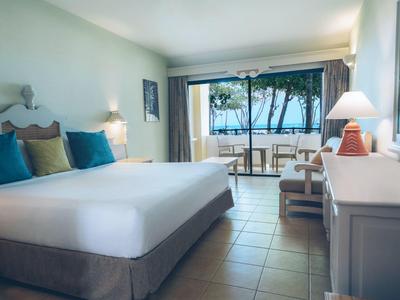 Helles Hotelzimmer mit großem Bett und Balkon mit Blick auf einen bewaldeten Bereich.