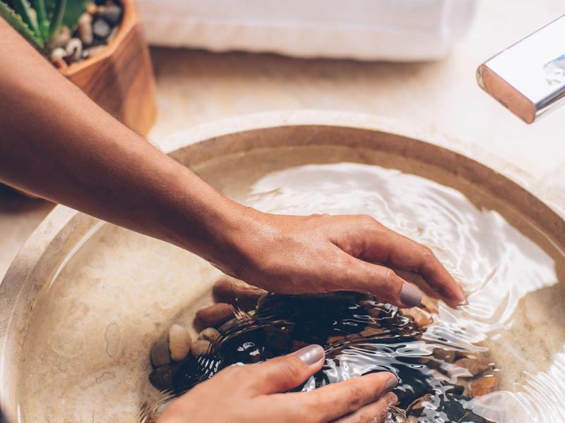 Hände tauchen in einen runden Steinwaschbecken mit Wasser und Steinen, Spa-Handtuch im Hintergrund