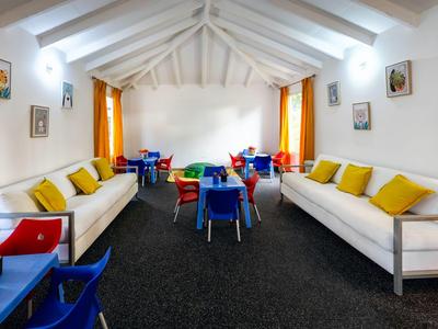 Heller Gruppenraum mit weißen Sofas, gelben Kissen und bunten Stühlen unter einem weißen Holzdach.