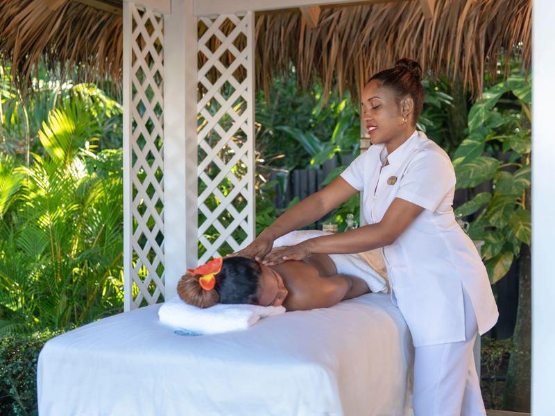 Eine Frau bekommt Massage im Freien unter einem Strohdach mit grünen Pflanzen im Hintergrund.