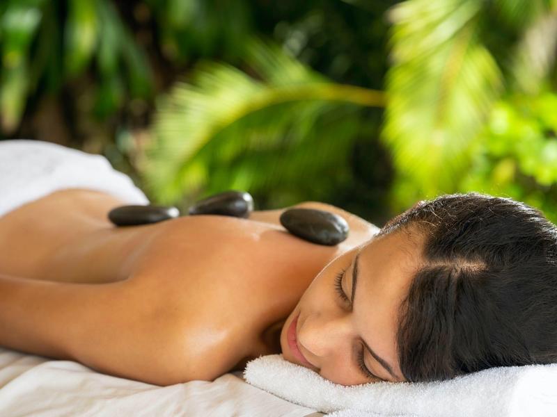 Frau liegt entspannt auf Bauch mit warmen, glatten Steinen auf dem Rücken in grünem Garten.