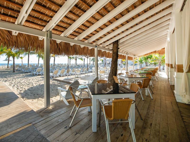 Offene Strandbar mit Holzdielen, Plastikstühlen und Tischen unter Strohdach, Palmen im Hintergrund.