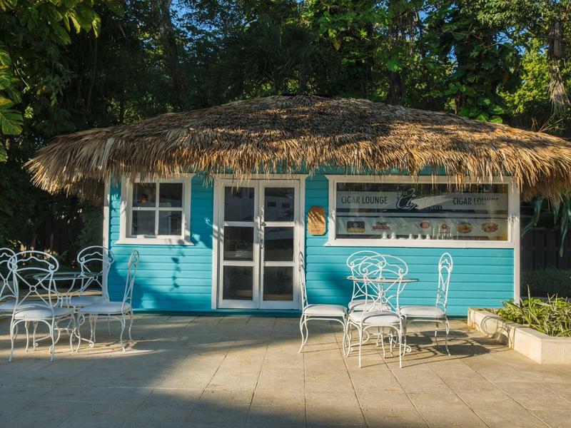 Kleines blaues Haus mit Strohdach und weißen Metallstühlen auf gepflastertem Boden.