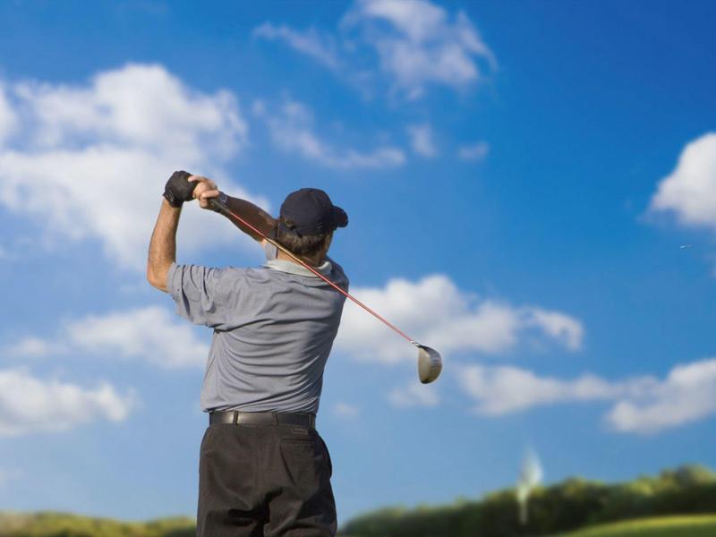 Mann in grauem Hemd und schwarzer Hose schlägt Golfball vor blauem Himmel mit Wolken.