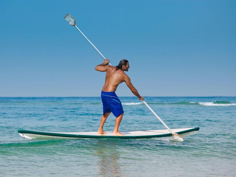 Mann in blauer Badehose steht auf Paddleboard und paddelt im klaren Meerwasser bei klarem Himmel.