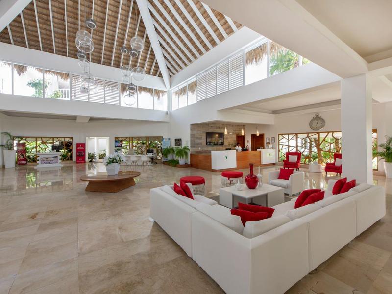 Großes helles Wohnzimmer mit weißen Sofas, roten Kissen und hohen Fenstern unter offenem Holztragwerk.