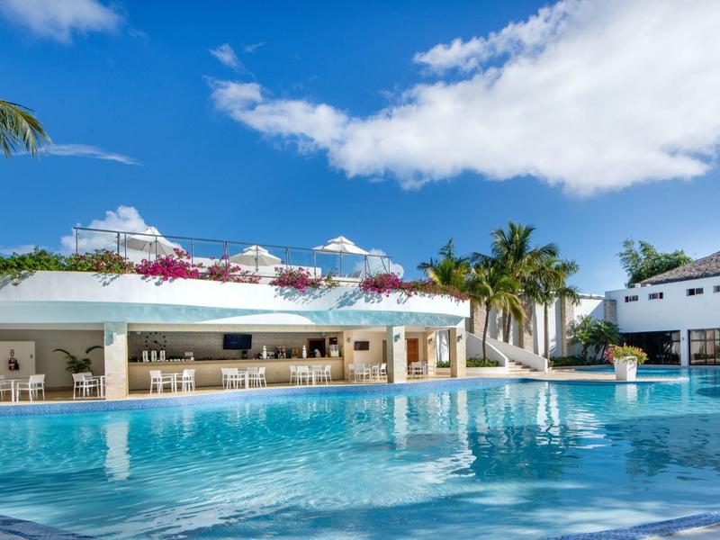Moderner Poolbereich mit klarem Wasser, Palmen und weißen Gebäuden unter blauem Himmel.