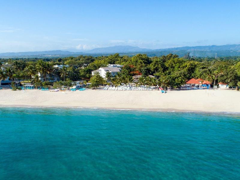 Klarer blauer Ozean vor weißem Sandstrand mit Palmen und Gebäuden im Hintergrund.