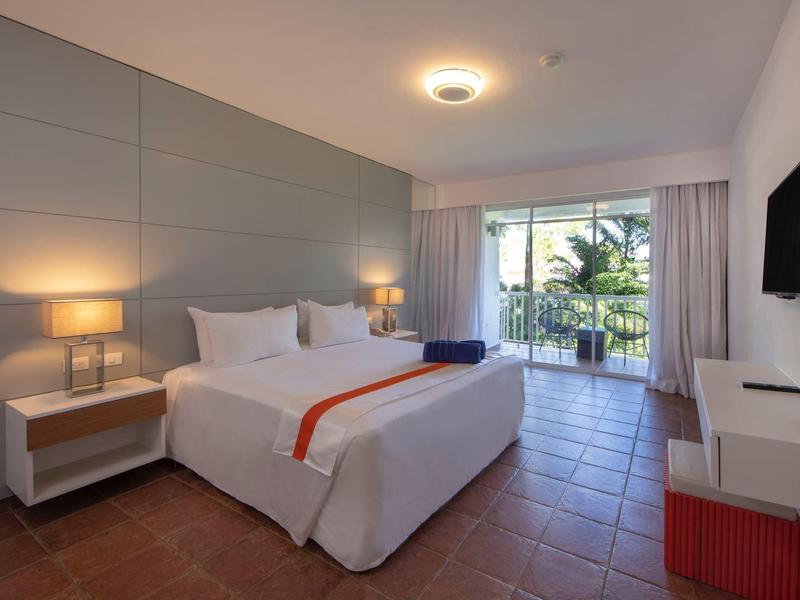 Hell eingerichtetes Schlafzimmer mit großem Bett, zwei Nachttischen, Balkon und Flachbildfernseher an der Wand.