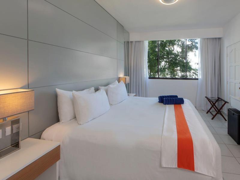 Modernes, schlichtes Schlafzimmer mit großem Bett, weißen Wänden, Fenster und sanfter Beleuchtung.