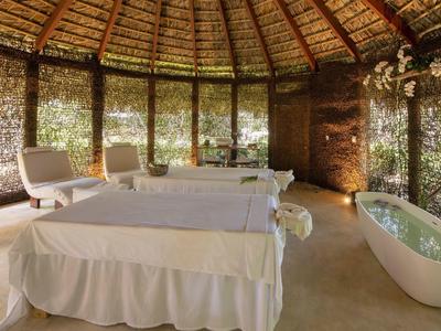 Innenraum einer Massagehütte mit Strohdach, zwei Massagetischen, weißem Wannne und Stühlen.
