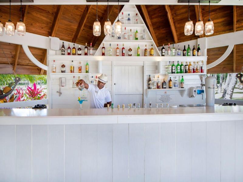 Moderner, heller Barbereich mit Holzdecke, weißen Möbeln, Flaschenregal und Barkeeper hinter Tresen.