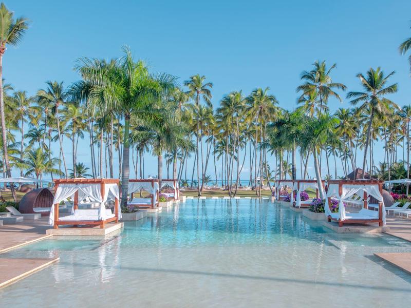 Großer Pool mit weißen Liegen und Palmen, blauer Himmel im Hintergrund.
