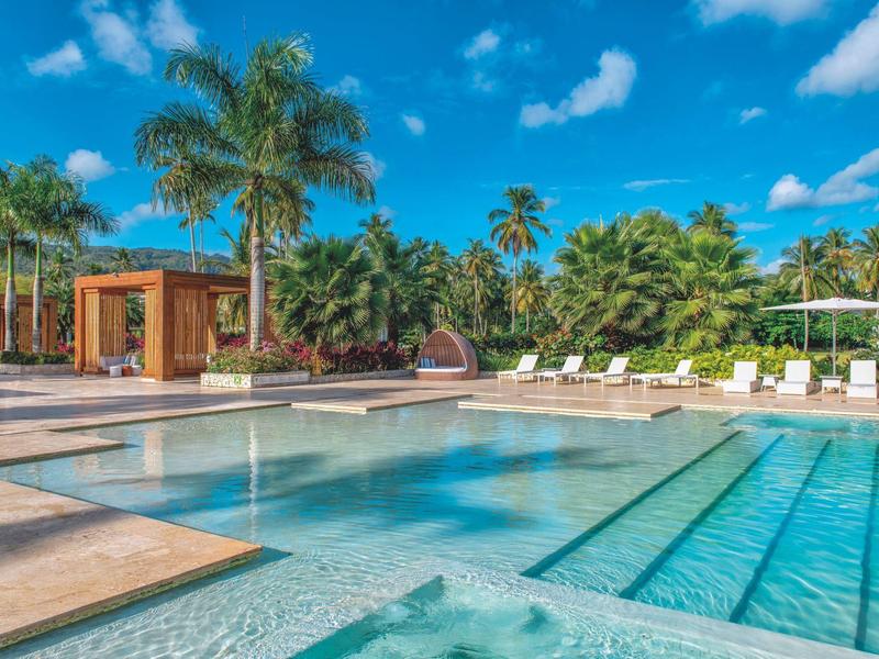 Großer Pool mit klar blauem Wasser, umgeben von Palmen und weißem Liegestuhlbereich.