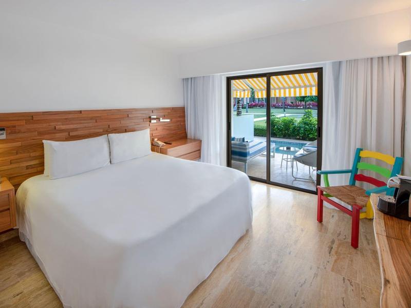 Helles Hotelzimmer mit Doppelbett, Holzmöbeln, Sitzbereich und Terrasse mit Blick auf Pool und Garten.