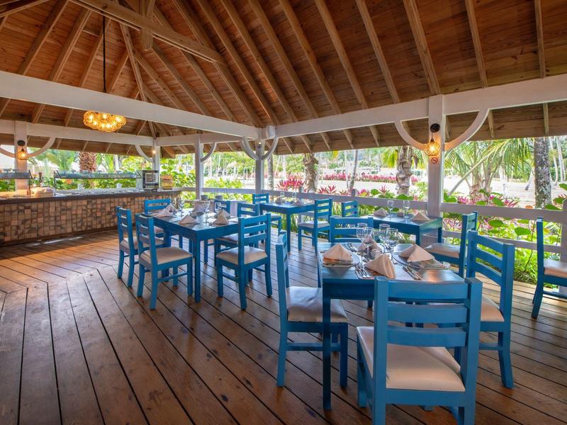 Offenes Restaurant mit hölzernem Boden, blauen Stühlen und Tischen, umgeben von grüner Natur.