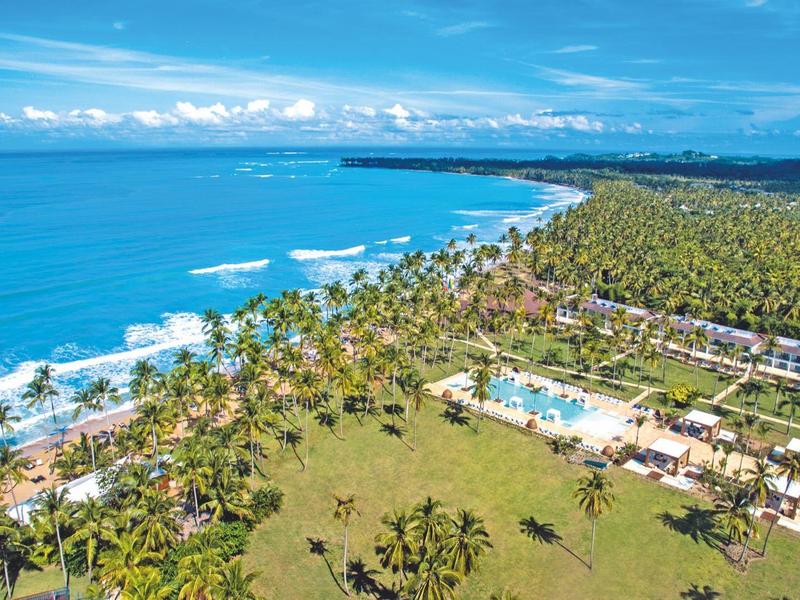 Langer Sandstrand mit Palmen, blaues Meer, Hotelanlage und grüner Vegetation bei Sonnenschein.